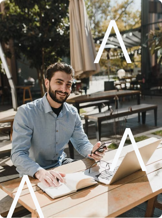 Un homme assis à une table en extérieur, un livre ouvert et un ordinateur portable devant lui. Il tient un téléphone. 