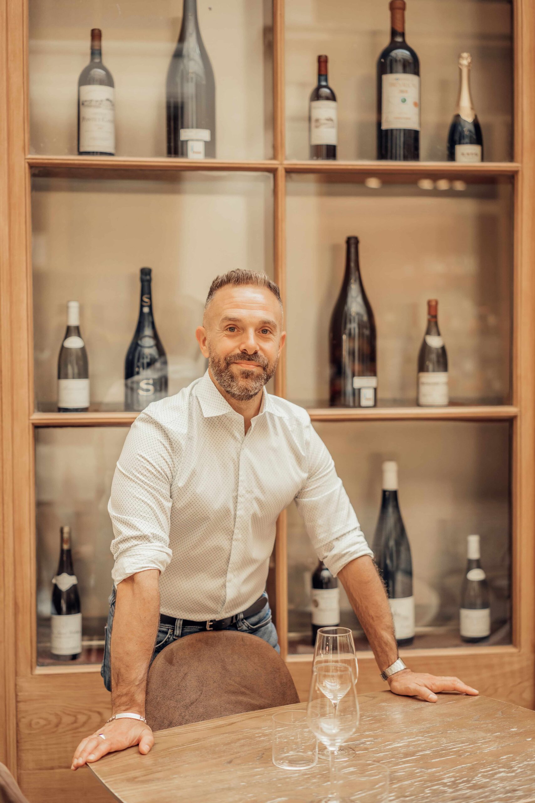 Sommelier devant des rayonnages de vin. Il a les mains sur une table en bois.